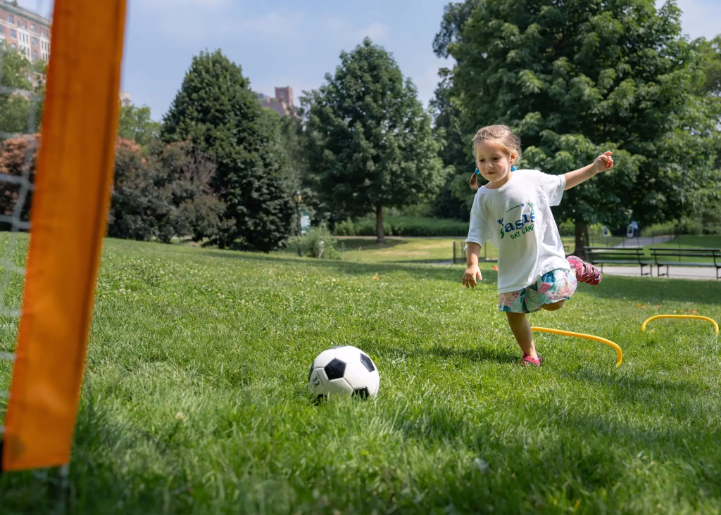 Young girl in a white "Oasis Day Camp" shirt practices soccer in a grassy park on a sunny day, aiming near an orange goal post.
